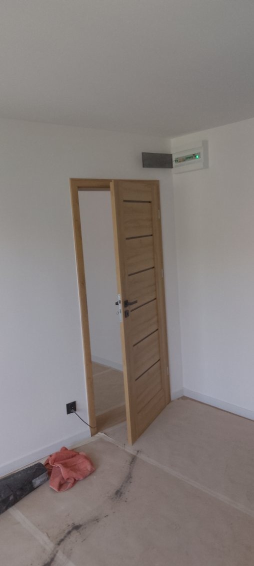 Jasne, drewniane drzwi wewnętrzne z poziomymi frezami, częściowo otwarte w białym pomieszczeniu. Widoczna skrzynka elektryczna i fragment podłogi w trakcie prac wykończeniowych.