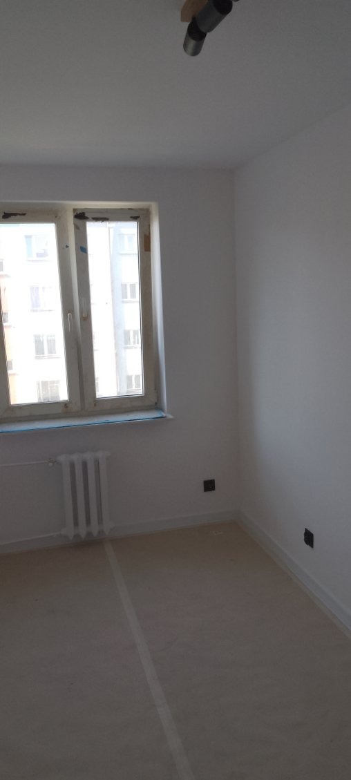 Jasne, świeżo pomalowane wnętrze pokoju z oknem i grzejnikiem. Widoczne gładkie ściany, przygotowane do dalszego wykończenia. Minimalistyczny styl, neutralna kolorystyka.