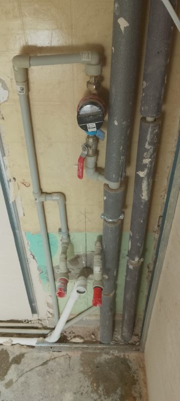 Szare rury z zaworami i licznikiem, instalacja wodna na tle ściany z resztkami tapety i surowego betonu. Widoczne łączenia i mocowania rur.