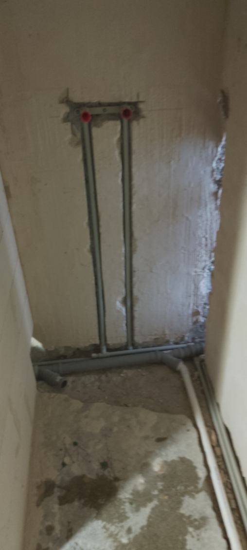 Szare rury instalacji wodnej w bruzdach ściany, z czerwonymi zaślepkami. Widoczne fragmenty podłogi i ścian w surowym stanie, w trakcie prac remontowych.