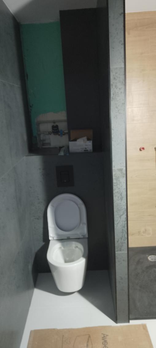 Wnętrze łazienki w trakcie remontu. Widoczna podwieszana toaleta, szare płytki, fragment ściany z instalacją i karton na podłodze. Ujęcie z góry.