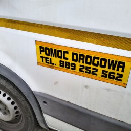 Sławomir Wojciechowski - Fragment boku białego samochodu dostawczego z żółtym pasem odblaskowym i naklejką z czarnym napisem 'POMOC DROGOWA TEL. 889 252 562', widoczne koło.