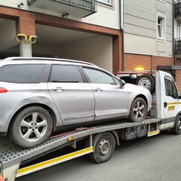 Sławomir Wojciechowski - Srebrny samochód osobowy typu kombi zabezpieczony pasami na platformie białej lawety z włączonym pomarańczowym światłem ostrzegawczym na tle budynku mieszkalnego.