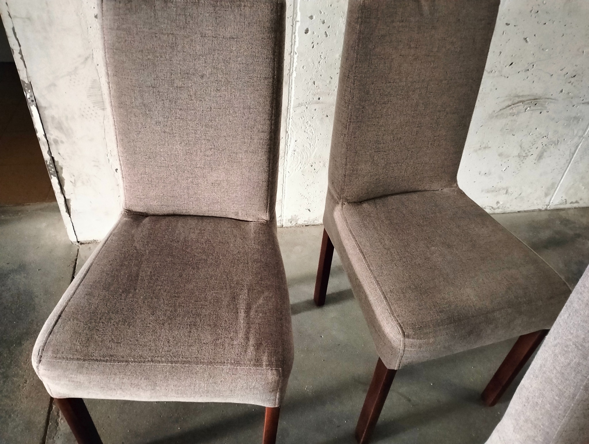 Dwa krzesła tapicerowane brązową tkaniną, z drewnianymi nogami, sfotografowane na tle surowej, betonowej ściany i posadzki.