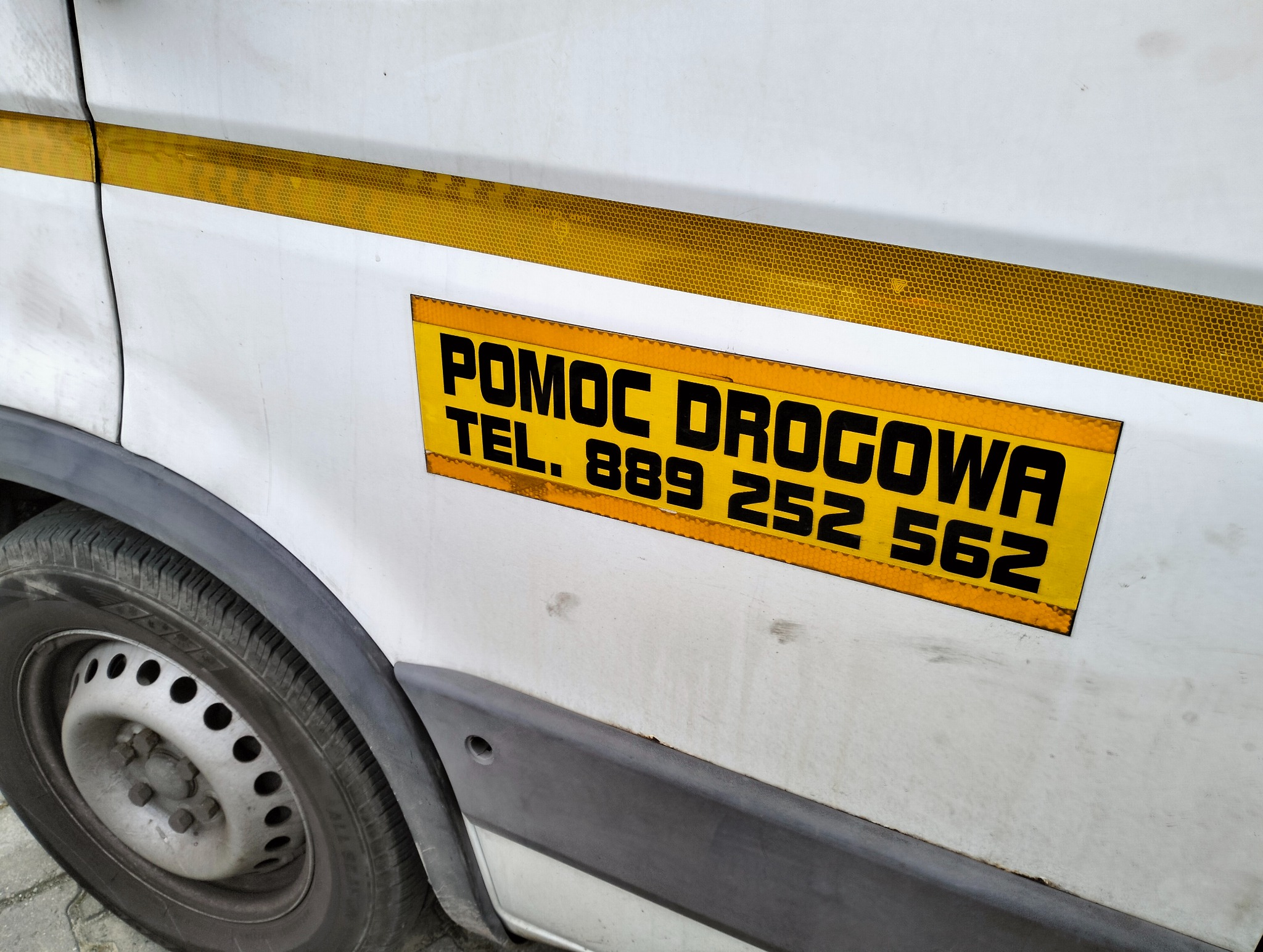 Fragment boku białego samochodu dostawczego z żółtym pasem odblaskowym i naklejką z czarnym napisem 'POMOC DROGOWA TEL. 889 252 562', widoczne koło.