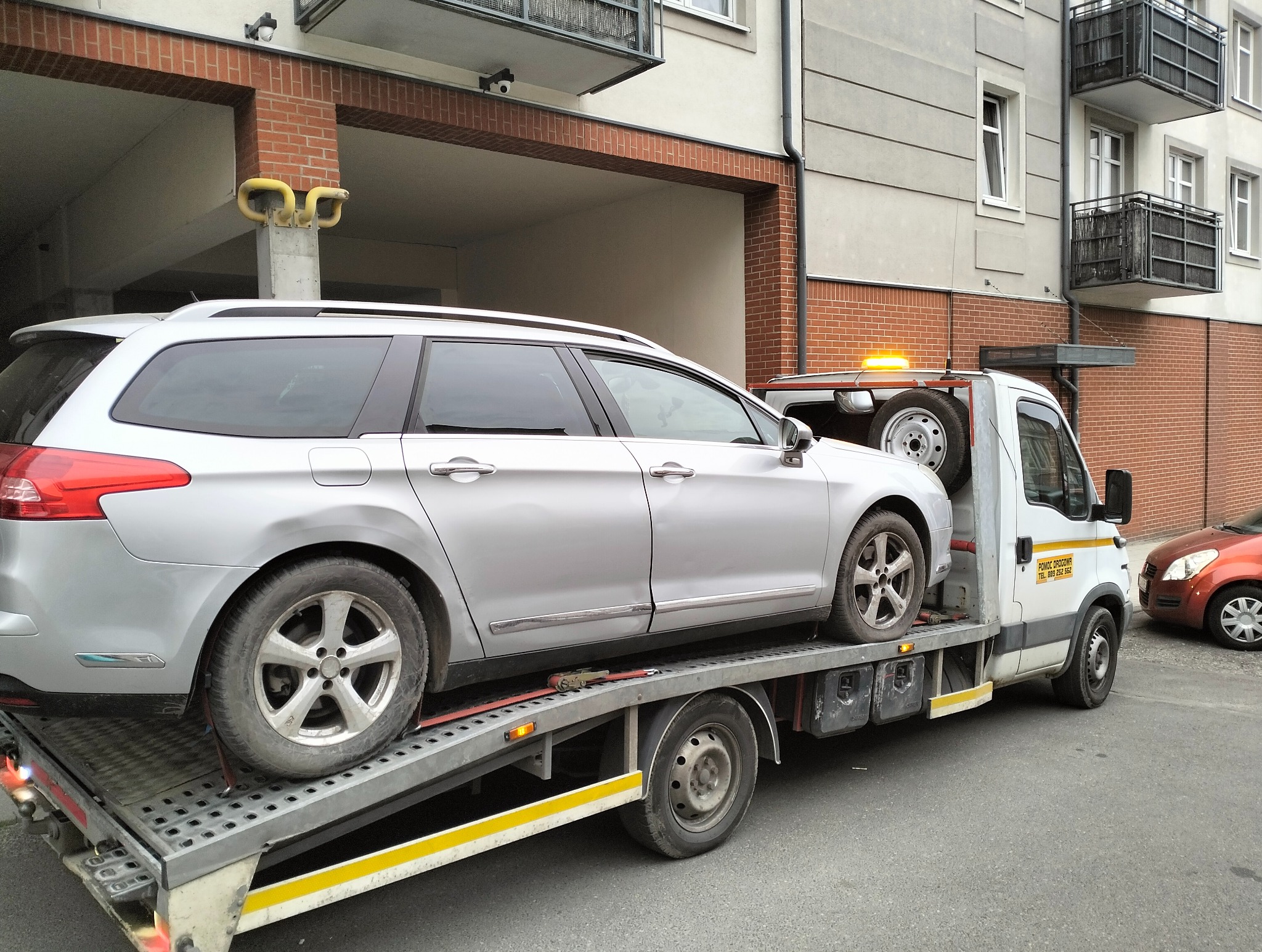 Srebrny samochód osobowy typu kombi zabezpieczony pasami na platformie białej lawety z włączonym pomarańczowym światłem ostrzegawczym na tle budynku mieszkalnego.