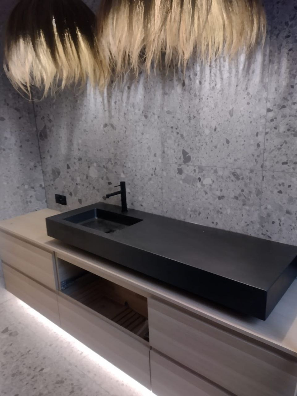 Nowoczesna łazienka: czarna, prostokątna umywalka nablatowa z czarną baterią na szafce z podświetleniem LED, ściana wykończona płytkami imitującymi lastryko, dekoracyjne lampy wiszące.