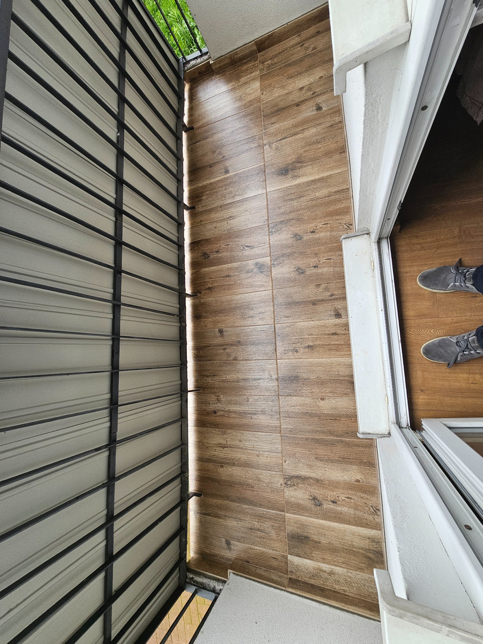Balkon z drewnopodobnymi płytkami gresowymi na podłodze, widok z góry, szare buty w kadrze, barierka balkonowa z metalowymi prętami.