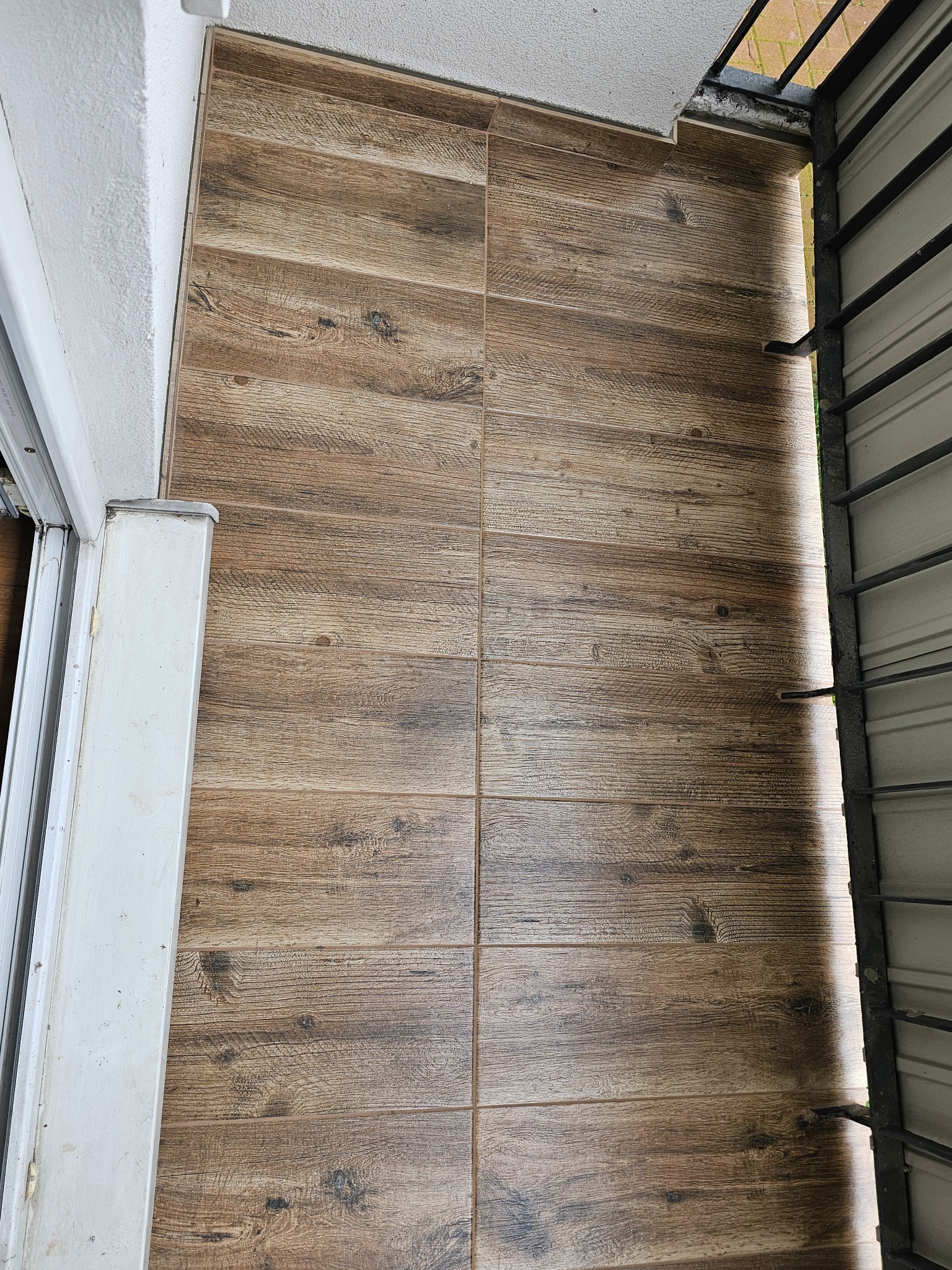 Ułożona podłoga z płytek imitujących drewno na balkonie, widok z góry, drzwi balkonowe i barierka z metalowych prętów po bokach.