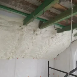 Poddasze z drewnianą konstrukcją dachu, pokryte grubą warstwą białej pianki izolacyjnej; widoczne elementy rusztowania i folia ochronna na ścianach.