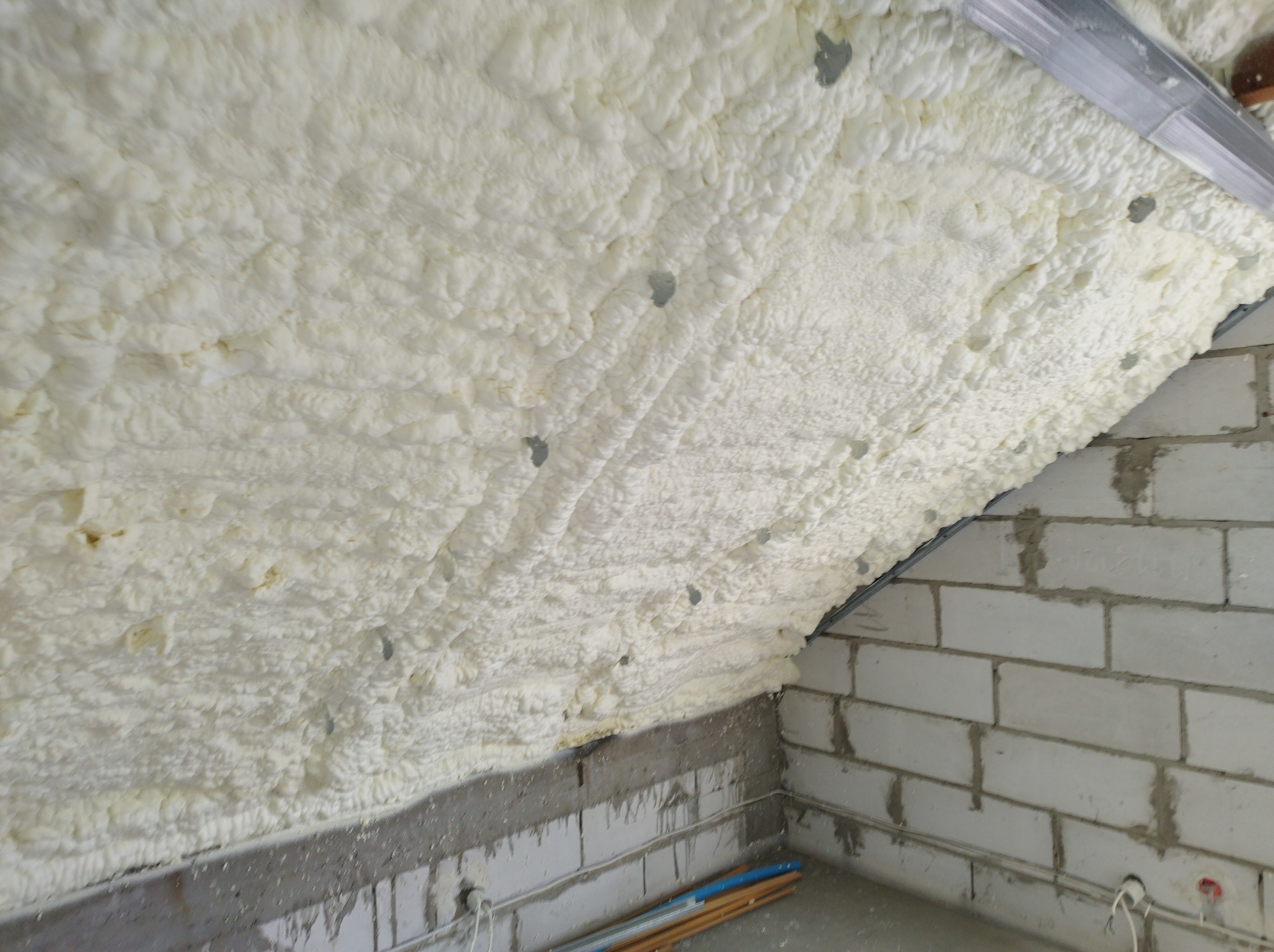 Poddasze z bloków betonowych po natrysku jasną, gęstą pianą izolacyjną, widoczne metalowe profile i fragment podłogi z rurami.