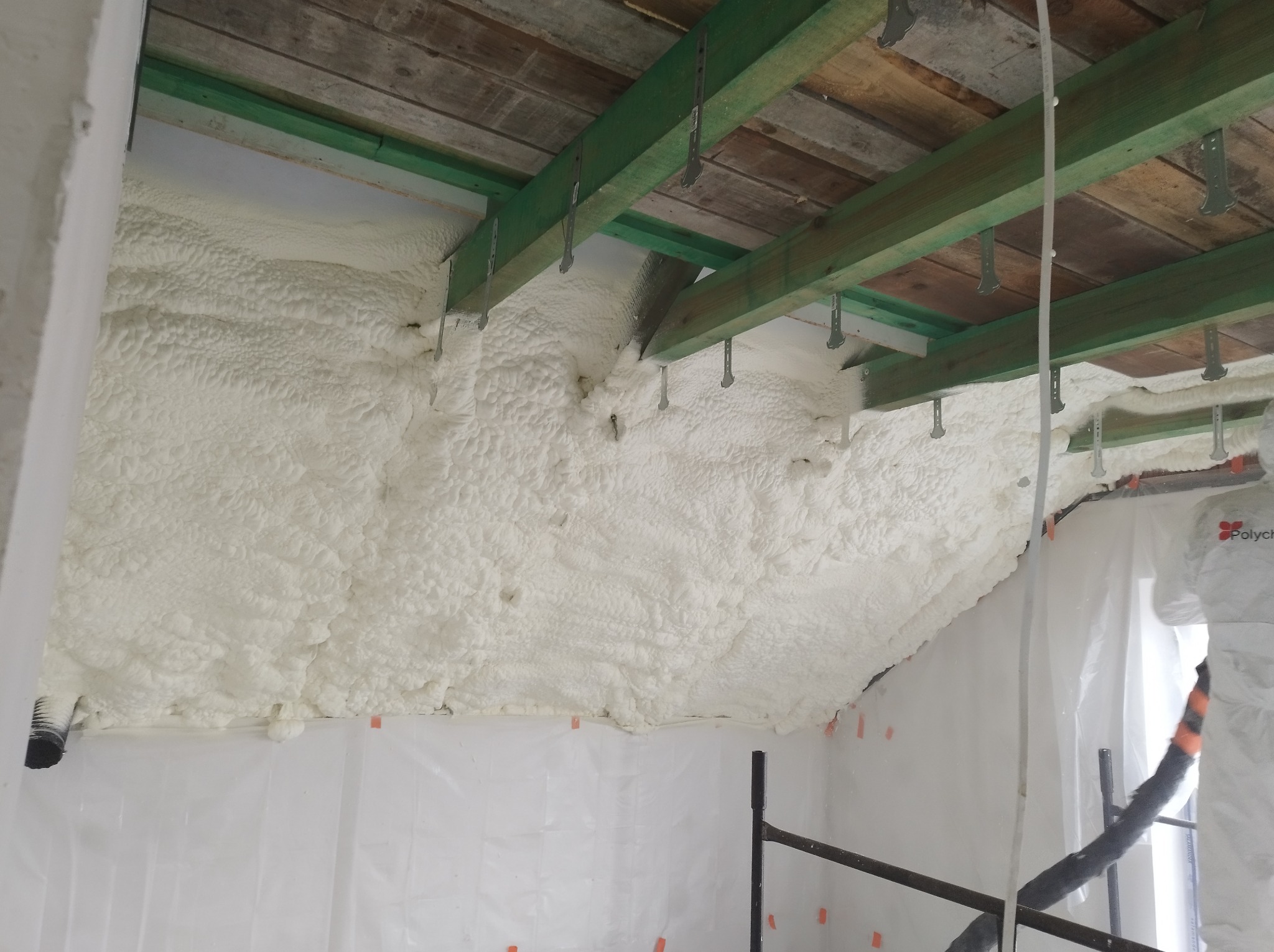 Poddasze z drewnianą konstrukcją dachu, pokryte grubą warstwą białej pianki izolacyjnej; widoczne elementy rusztowania i folia ochronna na ścianach.