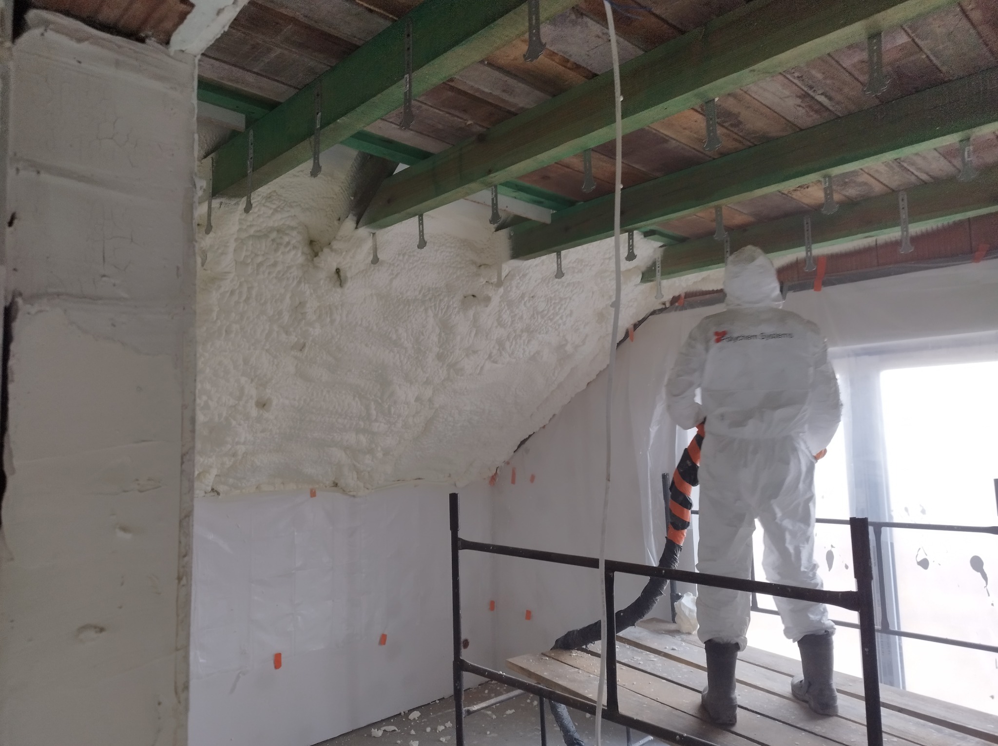 Pracownik w kombinezonie ochronnym aplikuje pianę izolacyjną na poddaszu, widoczne drewniane elementy konstrukcji dachu i rusztowanie.