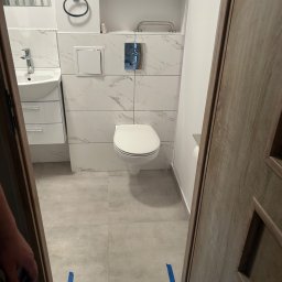 KONRAD BANACH Remonty i wykończenia wnętrz - Wykończona łazienka z białymi płytkami imitującymi marmur, wisząca toaleta, umywalka z szafką i drewniane drzwi z widoczną listwą przypodłogową zabezpieczoną niebieską taśmą malarską.