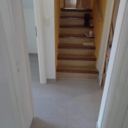 KONRAD BANACH Remonty i wykończenia wnętrz - Widok na wyremontowane schody z wykładziną dywanową i brązowymi obramowaniami, widoczne z korytarza z szarą podłogą z płytek; na schodach stoi czarny głośnik i biały kubek na tacy.