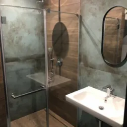 Nowoczesna kabina prysznicowa ze szklanymi drzwiami i deszczownicą, umywalka nablatowa z chromowaną baterią, lustro w czarnej ramie, płytki imitujące drewno i beton na ścianach, minimalistyczne...