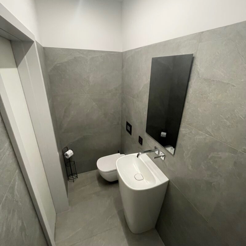 Nowoczesna, szara łazienka z wiszącą toaletą, wolnostojącą umywalką i czarnym lustrem, wykończona płytkami imitującymi kamień.