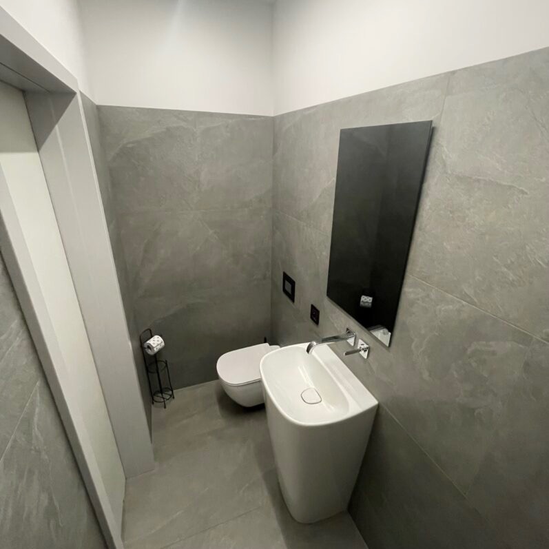 Nowoczesna łazienka w odcieniach szarości z wiszącą toaletą, umywalką wolnostojącą i czarnym lustrem na płytkach imitujących kamień.