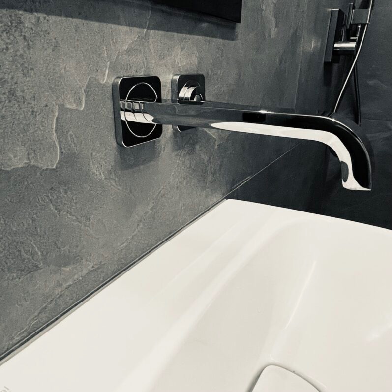 Bateria umywalkowa ścienna z chromowanym wykończeniem na tle szarej ściany z efektem kamienia, widoczna część białej umywalki.