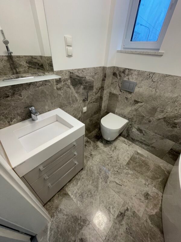 Nowoczesna łazienka z marmurowymi płytkami, wiszącą toaletą i szafką z umywalką w szarym odcieniu, oświetlona naturalnym światłem z okna dachowego.