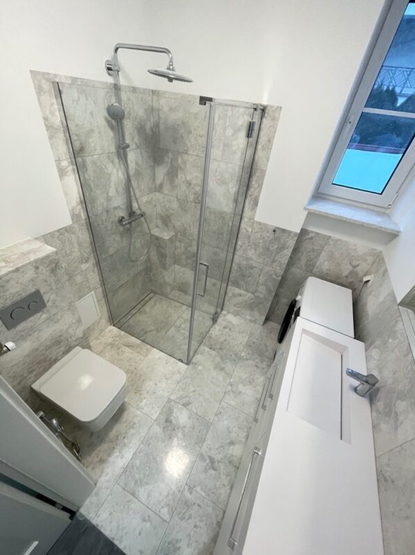 Nowoczesna łazienka na poddaszu z kabiną prysznicową, toaletą podwieszaną i białą szafką z umywalką, wykończona szarą marmurową płytką.