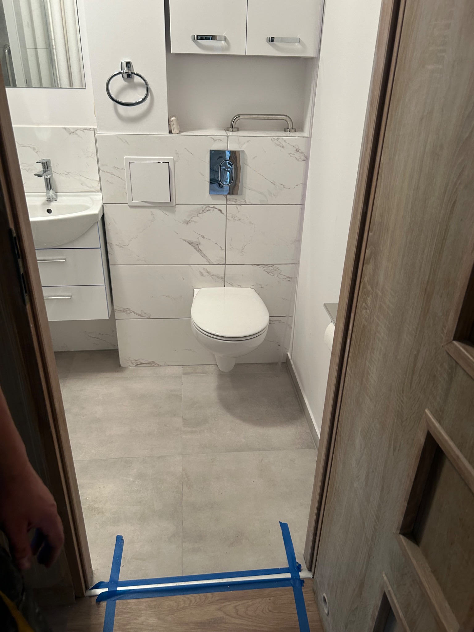 Wykończona łazienka z białymi płytkami imitującymi marmur, wisząca toaleta, umywalka z szafką i drewniane drzwi z widoczną listwą przypodłogową zabezpieczoną niebieską taśmą malarską.