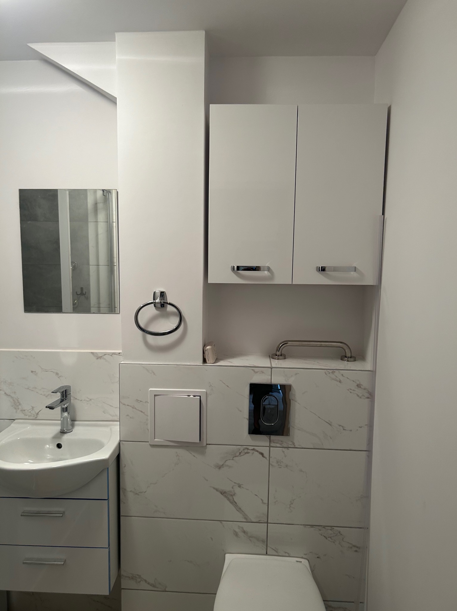 Nowoczesna, minimalistyczna łazienka z białymi szafkami, umywalką z szafką, lustrem i marmurowymi płytkami na ścianach. Widoczny fragment toalety i uchwyt dla niepełnosprawnych.