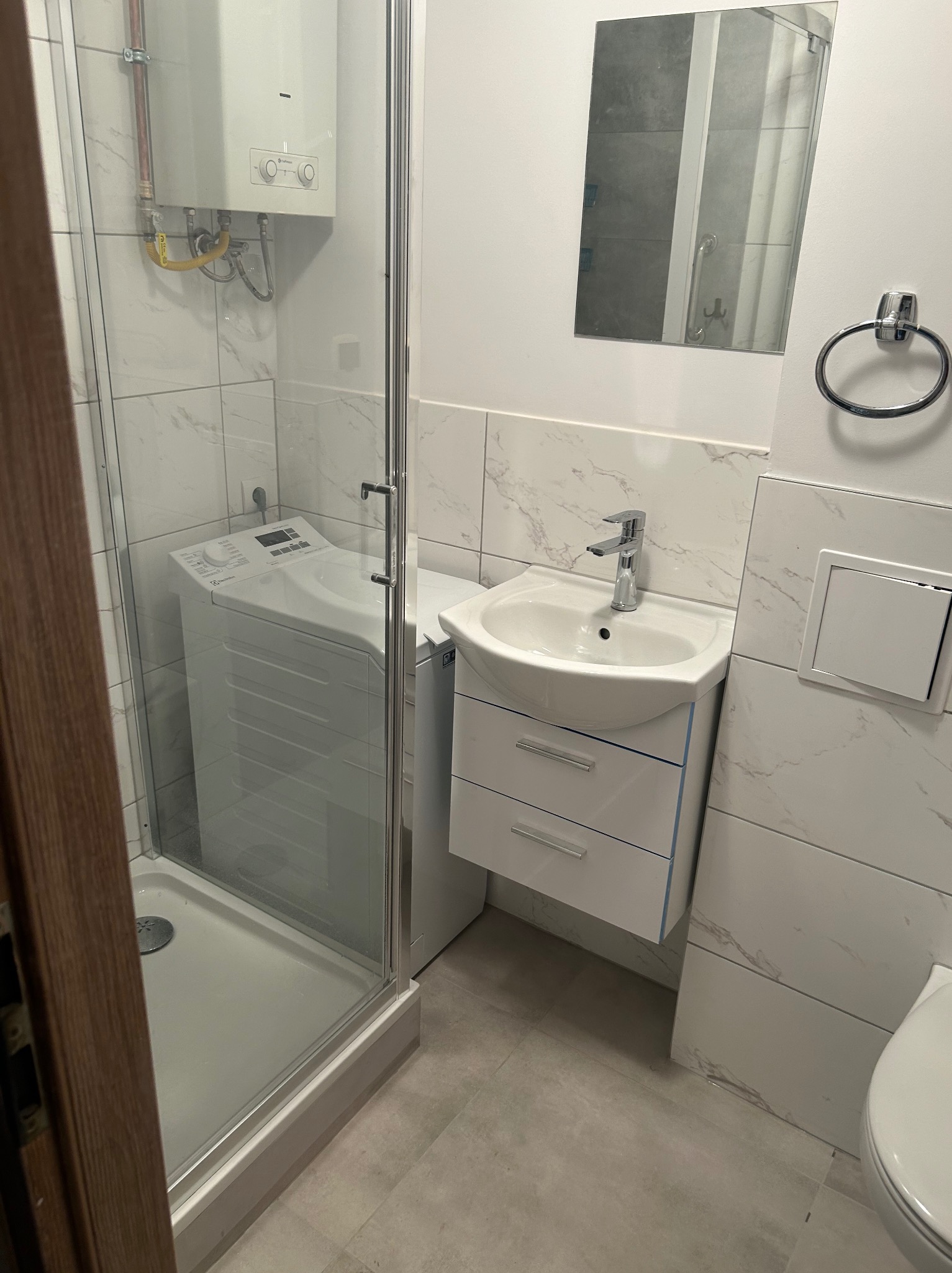 Nowoczesna łazienka z kabiną prysznicową, umywalką z szafką, lustrem, wiszącą toaletą i pralką w kabinie prysznicowej, wykończona białymi płytkami imitującymi marmur.