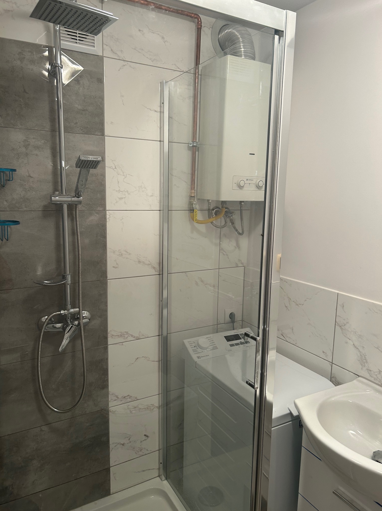 Nowoczesna kabina prysznicowa z deszczownicą i baterią, obok piecyk gazowy, pralka i umywalka w łazience wykończonej płytkami imitującymi marmur.