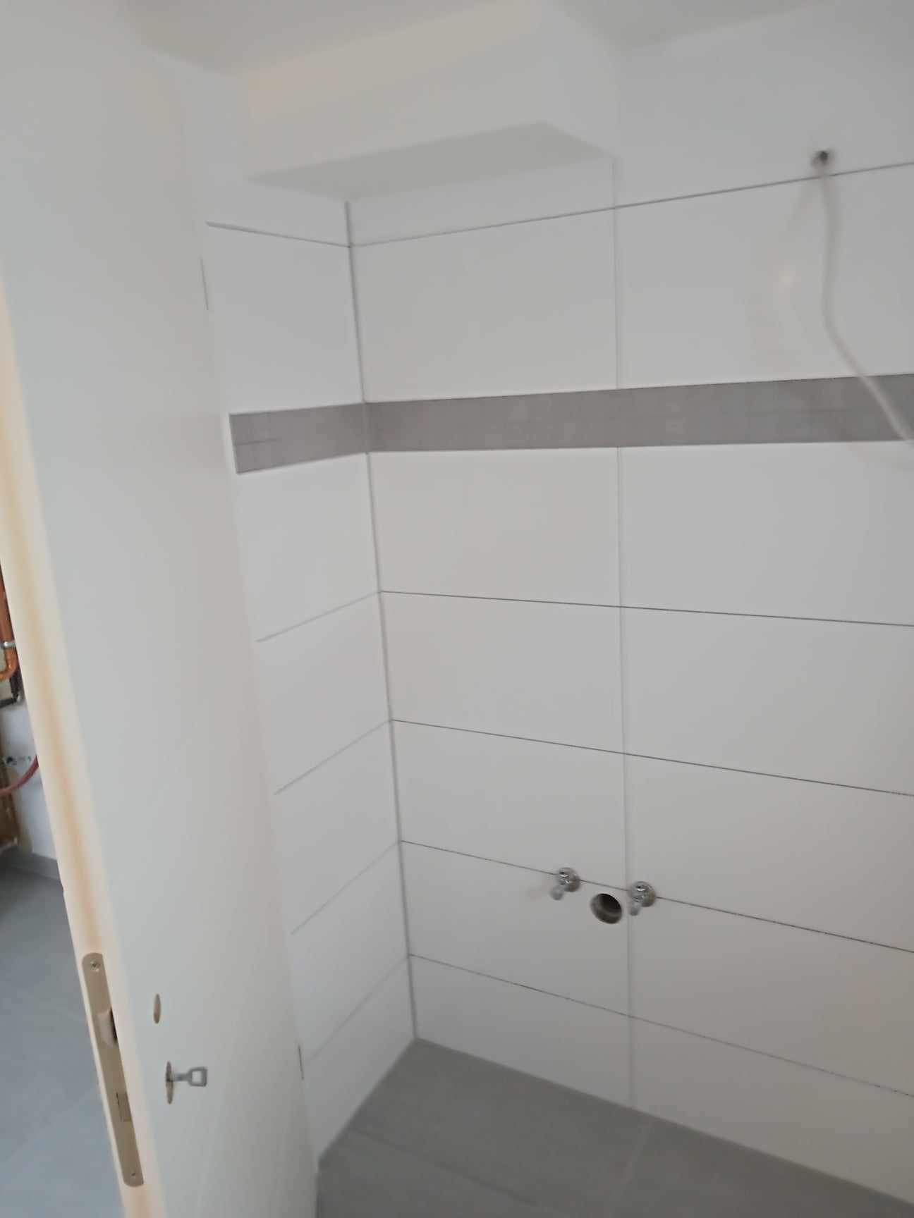 Świeżo wykafelkowana, jasna łazienka z szarym pasem dekoracyjnym i przygotowanymi przyłączami wodnymi, widok zza uchylonych drzwi.