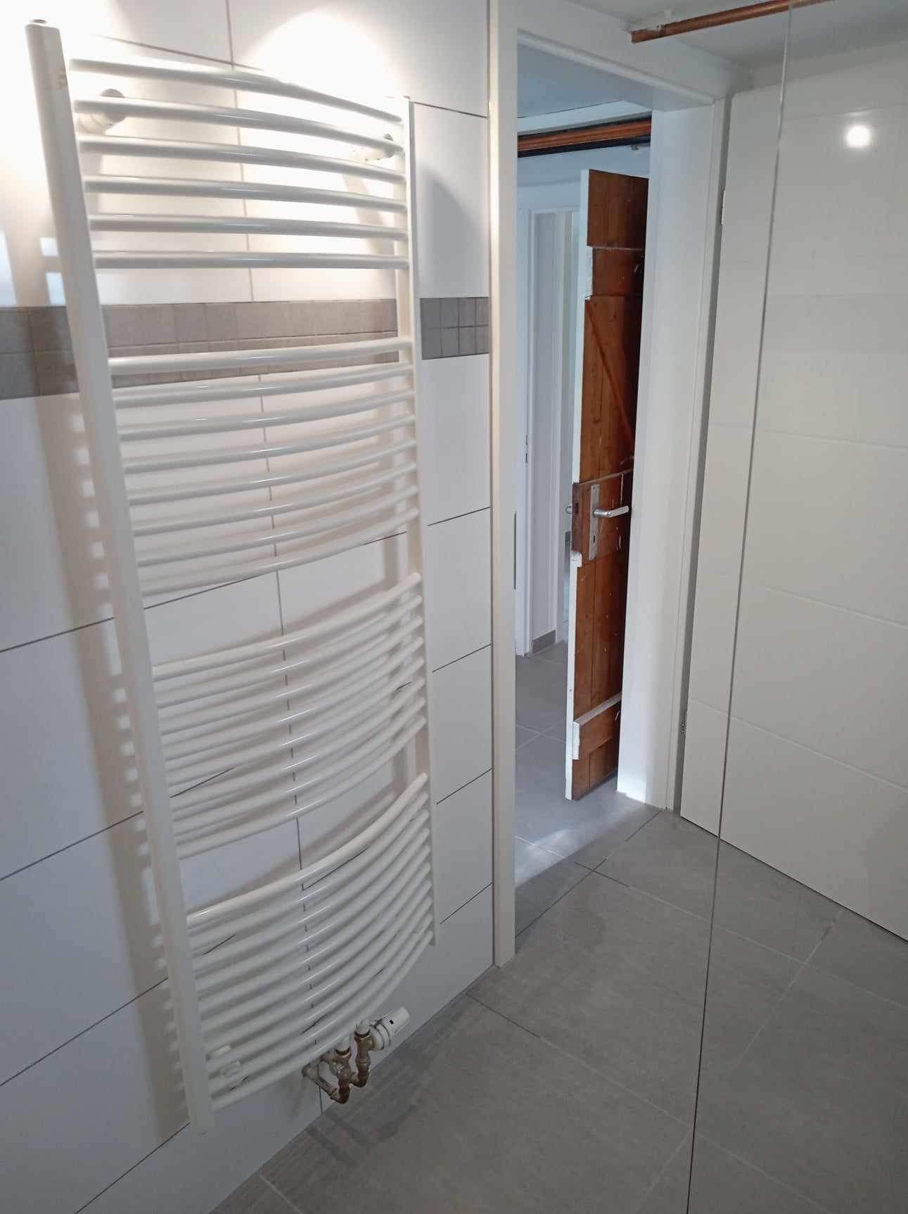 Biały, żeberkowy grzejnik łazienkowy na ścianie z białych płytek, widoczne przyłącza i termostat, szara podłoga i otwarte drzwi do innego pomieszczenia z drewnianymi drzwiami.