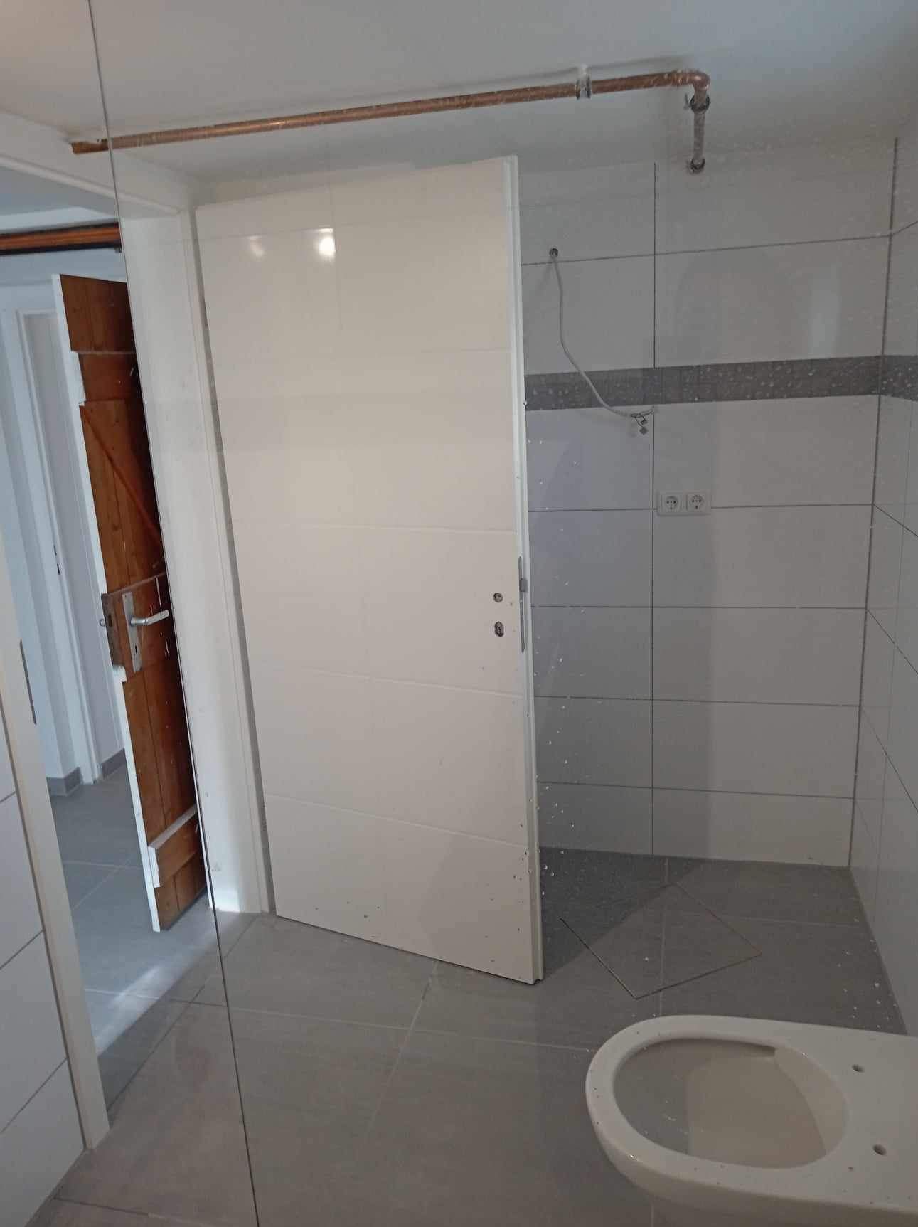 Wnętrze łazienki w trakcie prac: widoczne białe płytki, szara podłoga, częściowo zamontowana toaleta, białe drzwi z metalową klamką i miedziana rura pod sufitem.