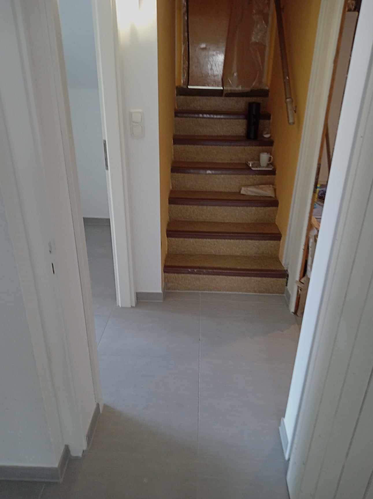 Widok na wyremontowane schody z wykładziną dywanową i brązowymi obramowaniami, widoczne z korytarza z szarą podłogą z płytek; na schodach stoi czarny głośnik i biały kubek na tacy.