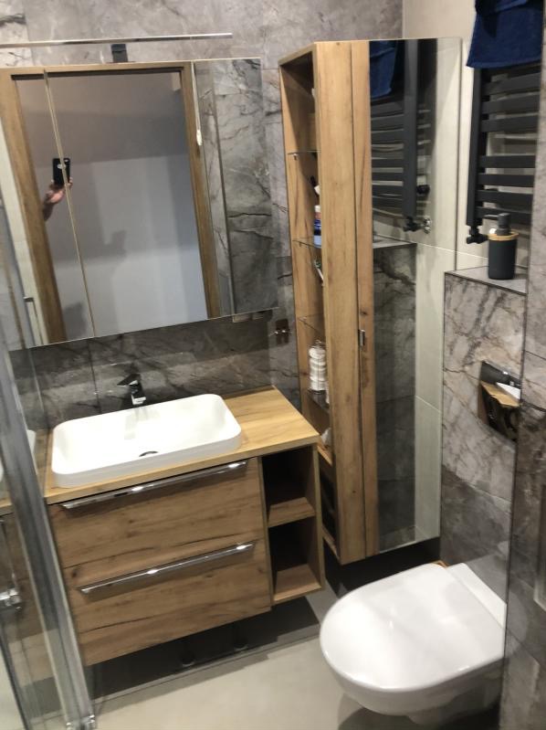 Nowoczesna łazienka z umywalką nablatową, szafką z drewnianym wykończeniem, wysoką szafką i toaletą w kolorze białym, na tle szarych płytek imitujących marmur.