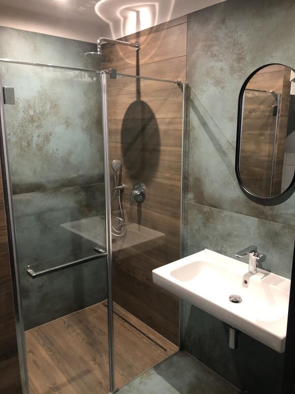 Nowoczesna kabina prysznicowa ze szklanymi drzwiami i deszczownicą, umywalka nablatowa z chromowaną baterią, lustro w czarnej ramie, płytki imitujące drewno i beton na ścianach, minimalistyczne...