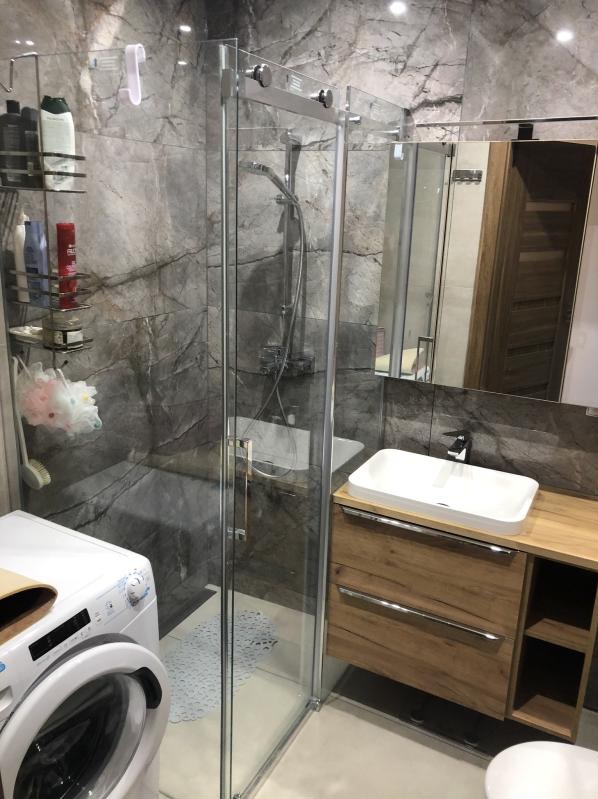 Nowoczesna łazienka w szarościach z kabiną prysznicową ze szklanymi drzwiami, umywalką nablatową na drewnianej szafce i pralką z przodu.