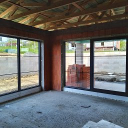 F.U.H. MIGACZ MAREK - Wnętrze surowego domu w trakcie budowy z zamontowanymi oknami z czarnymi ramami, widoczne ułożone cegły obok okna, surowy beton na podłodze i widok na ogród przez okna.