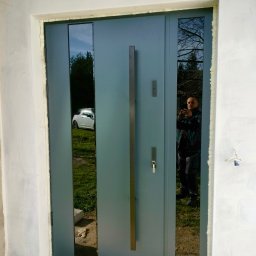 F.U.H. MIGACZ MAREK - Szare drzwi wejściowe z długim uchwytem i szybami bocznymi, widoczny montaż z pianką uszczelniającą, odbicie osoby robiącej zdjęcie w szybie.