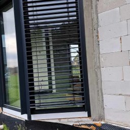 F.U.H. MIGACZ MAREK - Ciemne żaluzje poziome zamontowane na oknie budynku w trakcie budowy, widoczny surowy mur i fragment trawnika.