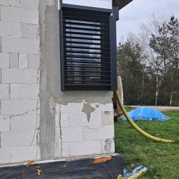 F.U.H. MIGACZ MAREK - Zamontowana czarna roleta zewnętrzna na oknie w surowym murze z bloczków, widoczny fragment dachu, żółty wąż i niebieska plandeka na trawie.