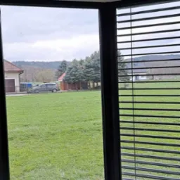 Wnętrze pokoju z narożnym oknem PCV z czarną ramą i zamontowanymi poziomymi żaluzjami, widok na trawnik, dom i zaparkowany samochód w pochmurny dzień.