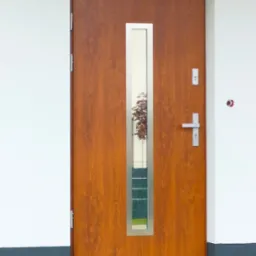 Brązowe drzwi wejściowe z pionowym, srebrnym przeszkleniem odbijającym drzewo, zamontowane w białej ścianie z szarą podmurówką.