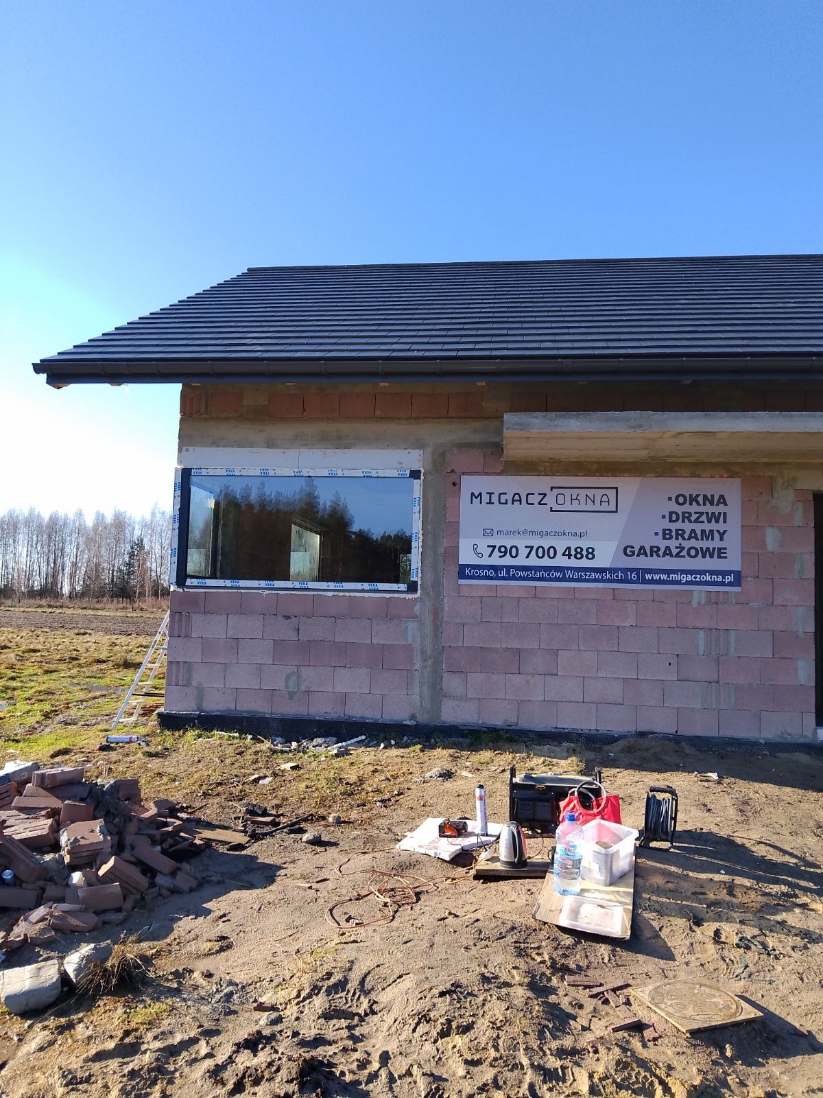 Montaż okna w surowym budynku z cegły, widoczny fragment dachu, narzędzia i materiały budowlane na ziemi, tablica reklamowa firmy.