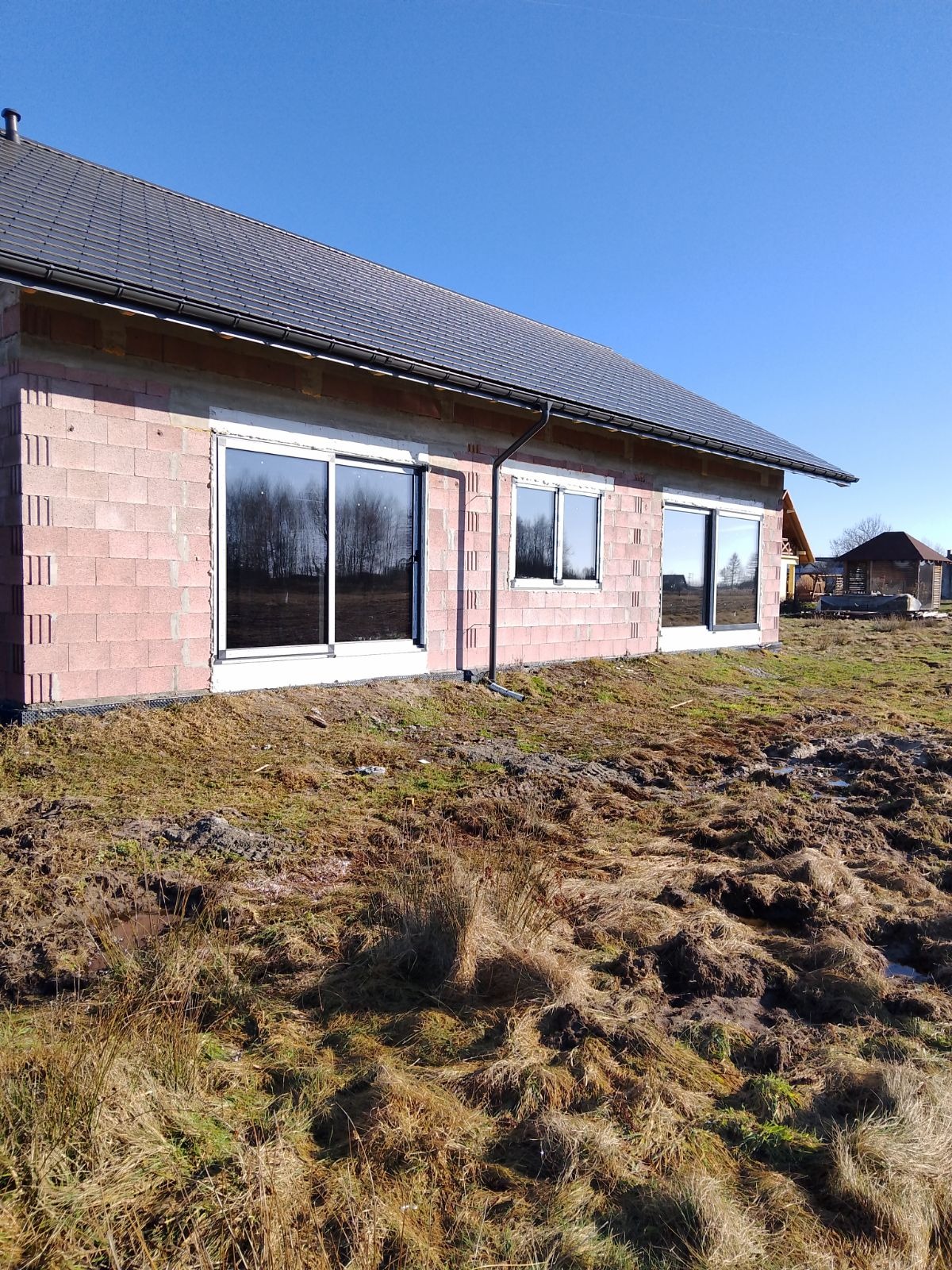 Surowy stan budynku z zamontowanymi oknami PCV z białymi ramami, widoczne odbicie drzew w szybach, teren wokół budynku zarośnięty trawą i błotem.