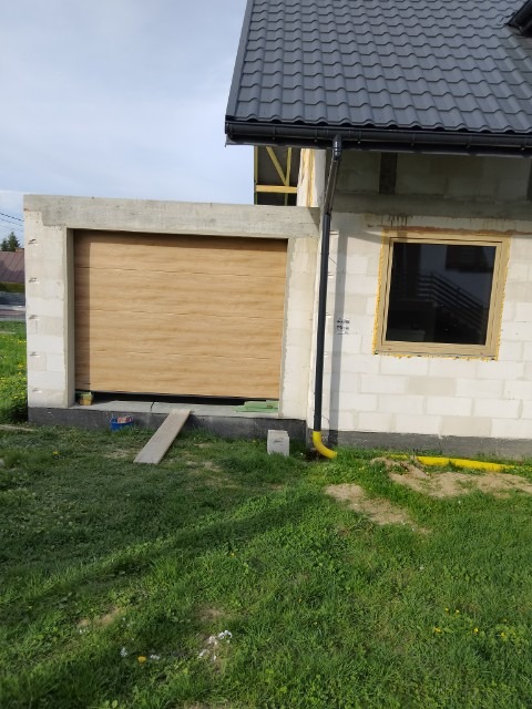 Jasnobrązowa brama garażowa segmentowa z poziomym wzorem drewna, zamontowana w surowym murze z betonu komórkowego, widok z zewnątrz na trawnik i fragment okna.