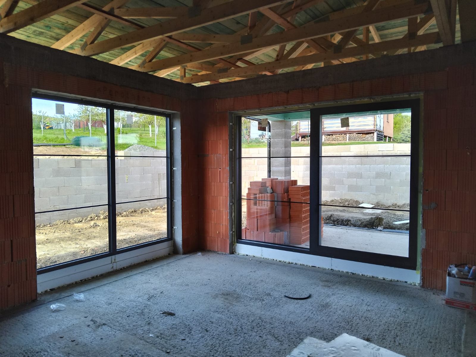 Wnętrze surowego domu w trakcie budowy z zamontowanymi oknami z czarnymi ramami, widoczne ułożone cegły obok okna, surowy beton na podłodze i widok na ogród przez okna.