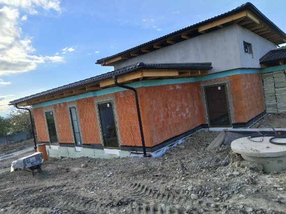 Budowa domu jednorodzinnego: widok na surową bryłę z zamontowanymi oknami i drzwiami, ściany z czerwonej cegły, ocieplenie, widoczne elementy drenażu i teren budowy.