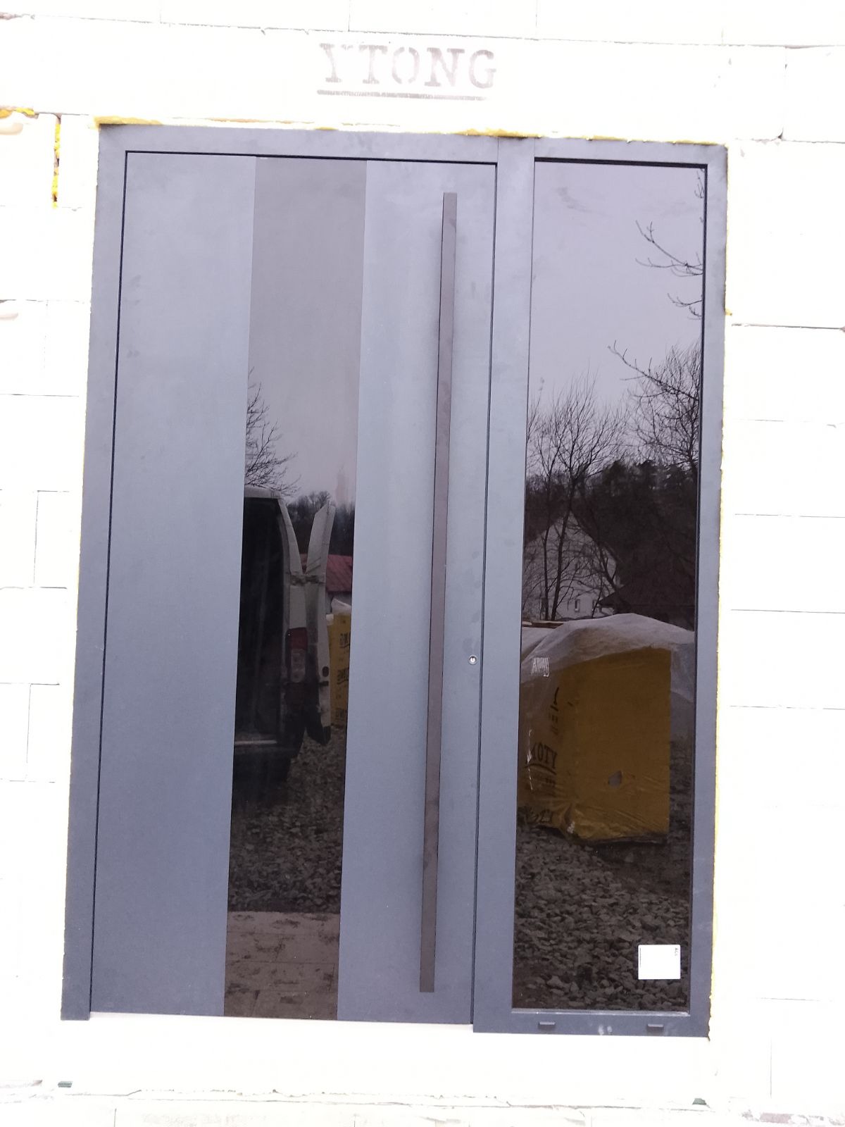 Szare drzwi wejściowe z długim, prostokątnym uchwytem, wbudowane w białą ścianę z bloków Ytong, z widocznym odbiciem samochodu dostawczego i materiałów budowlanych w szybie.