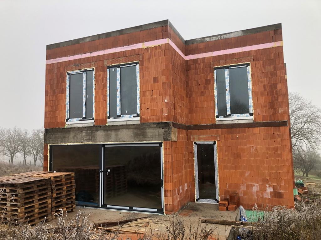 Surowy budynek w trakcie budowy z zamontowanymi oknami i drzwiami przesuwnymi, widoczne palety i cegły na zewnątrz.