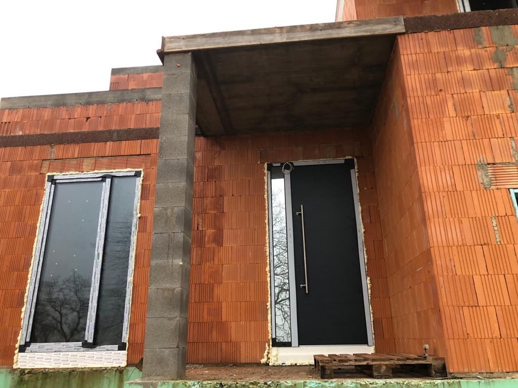 Surowy stan budowy: czarne drzwi wejściowe z długim, srebrnym uchwytem i okno zamontowane w ścianie z czerwonej cegły, widoczna piana montażowa, paleta leży przed drzwiami.